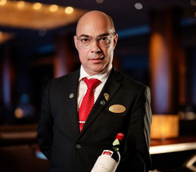 Luis Simoes in a black suit with a red tie and a gold nametag, holding a wine bottle